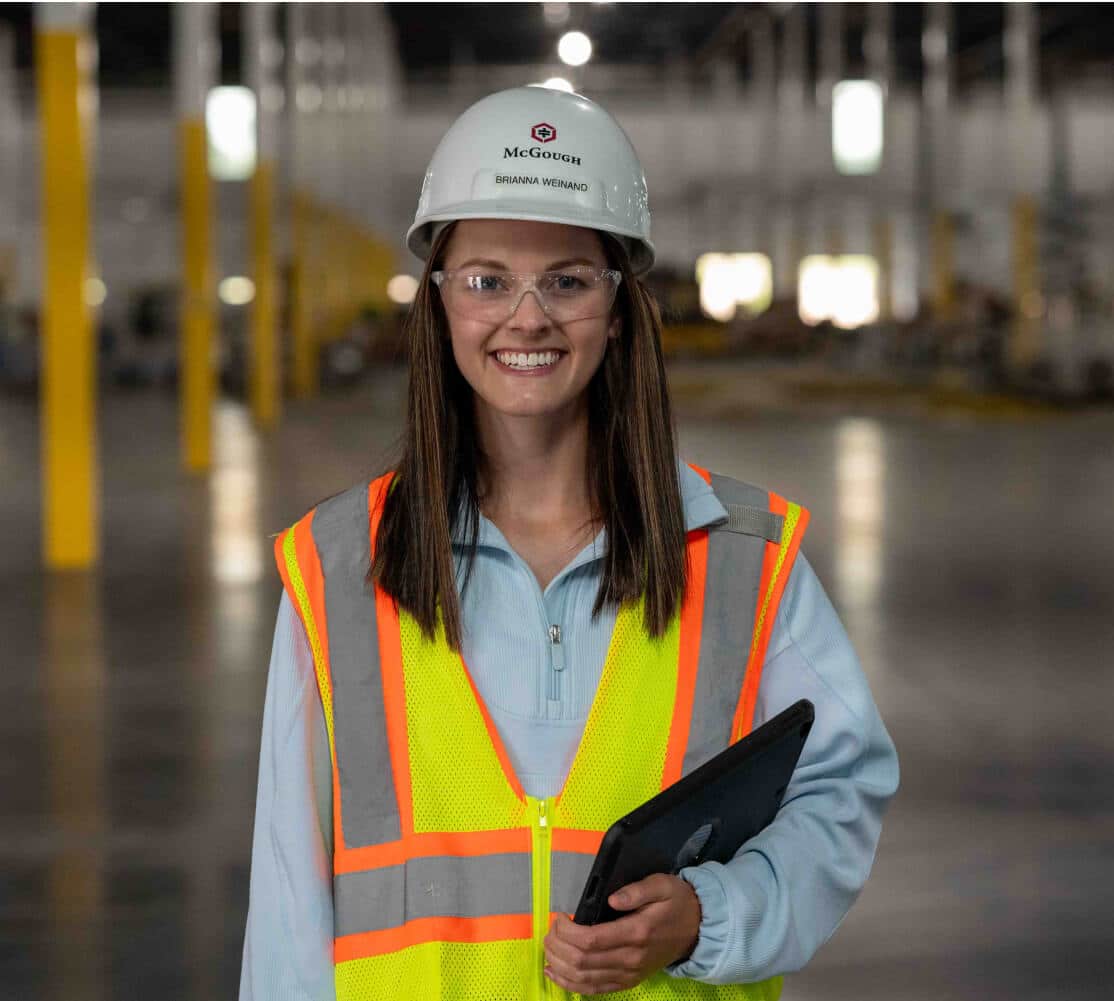 A smiling woman in McGough safety gear on a job site holding a computer