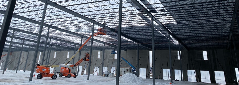 Interior image of a large industrial building under construction, with workers raising roof panels